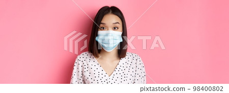 Covid-19, pandemic and lifestyle concept. Surprised asian woman in face mask raising eyebrows, staring at camera amazed, standing against pink background Covid-19, pandemic and lifestyle concept. Surprised asian woman in face mask raising eyebrows, staring at camera amazed, standing against pink background 98400802