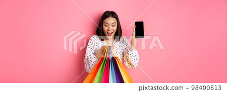 Attractive asian woman showing smartphone app and shopping bags, buying online via application, standing over pink background 98400813
