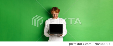 Happy young man with red hair, showing blank laptop screen and looking pleased, smiling while staring at computer, green background 98400927