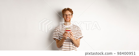 Holidays and celebration concept. Handsome redhead man in glasses holding birthday cake with candle, smiling and looking happy at camera Holidays and celebration concept. Handsome redhead man in glasses holding birthday cake with candle, smiling and looking happy at camera 98400951