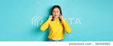 Tired and disappointed young asian woman unwilling to listen, shut ears with fingers and close eyes, standing in denial over blue background 98400960
