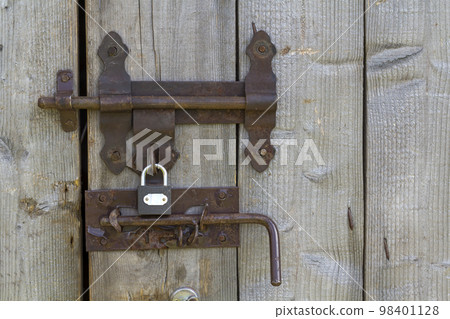 Close-up of lit by sun old rough made of wooden planks house door or barn gate with iron rusty slide bolt lock. Outdated technology, safety, security and protection from thieves concept. Close-up of lit by sun old rough made of wooden planks house door or barn gate with iron rusty slide bolt lock. Outdated technology, safety, security and protection from thieves concept. 98401128