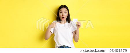 Young asian woman in casual white t-shirt showing plastic credit card and smiling at camera, yellow background 98401279
