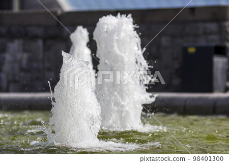 Close up of high mighty gush of cold clean sparkling transparent fresh water of a fountain. White foam and droplets on city background. 98401300