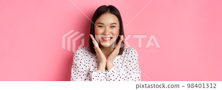 Beauty and skin care concept. Close-up of cute asian woman showing clean perfect face and smiling, looking happy at camera, standing over pink background 98401312