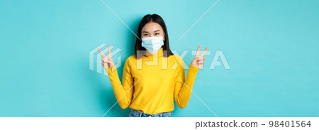 Covid-19, social distancing and pandemic concept. Kawaii asian woman showing peace signs and smiling happy, wearing medical mask from coronavirus disease, standing over blue background Covid-19, social distancing and pandemic concept. Kawaii asian woman showing peace signs and smiling happy, wearing medical mask from coronavirus disease, standing over blue background 98401564