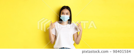 Covid-19, quarantine and social distancing concept. Young asian woman in white t-shirt, pointing at her medical mask from coronavirus, yellow background 98401584