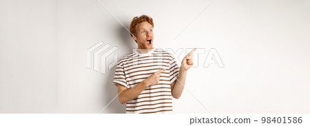 Check it out. Amazed redhead guy pointing and looking left at promotion banner, standing over white background Check it out. Amazed redhead guy pointing and looking left at promotion banner, standing over white background 98401586