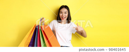 Cheerful asian woman going on shopping, pointing finger at paper bags and smiling, shopper having fun during discounts, yellow background Cheerful asian woman going on shopping, pointing finger at paper bags and smiling, shopper having fun during discounts, yellow background 98401645