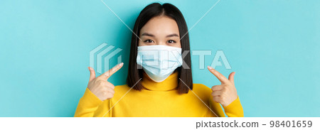 Covid-19, social distancing and pandemic concept. Close up of young asian woman using medical mask during coronavirus, standing over blue background Covid-19, social distancing and pandemic concept. Close up of young asian woman using medical mask during coronavirus, standing over blue background 98401659