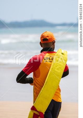 Lifeguard on duty, Walking on the sandy beach while carrying a rescue tube on the shoulder, lifeguard rear view photo. Lifeguard on duty, Walking on the sandy beach while carrying a rescue tube on the shoulder, lifeguard rear view photo. 98401834