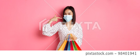 Covid-19, pandemic and lifestyle concept. Cute asian woman holding shopping bags, smiling and pointing at her face mask, standing over pink background 98401941