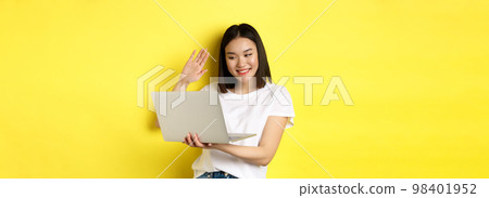 Happy asian woman video chat on laptop, waiving hand at computer camera and saying hello, standing over yellow background Happy asian woman video chat on laptop, waiving hand at computer camera and saying hello, standing over yellow background 98401952