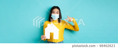 Covid-19, pandemic and real estate concept. Cheerful asian woman smiling in medical mask, showing paper house cutout, recommend agency for buying property, blue background 98402621
