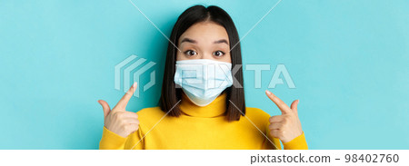 Covid-19, social distancing and pandemic concept. Close up of surprised asian girl pointing at her medical mask and looking amazed at camera, blue background 98402760