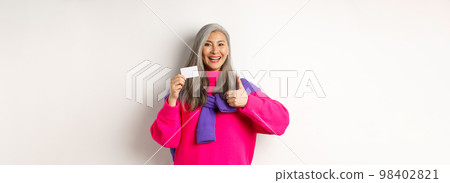 Shopping concept. Smiling asian middle-aged woman with grey hair showing plastic credit card and thumbs-up, recommending bank promotion, white background Shopping concept. Smiling asian middle-aged woman with grey hair showing plastic credit card and thumbs-up, recommending bank promotion, white background 98402821