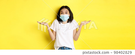 Covid-19, quarantine and social distancing concept. Clueless asian woman in medical mask shrugging shoulders, spread hands sideways clueless, know nothing, yellow background Covid-19, quarantine and social distancing concept. Clueless asian woman in medical mask shrugging shoulders, spread hands sideways clueless, know nothing, yellow background 98402829