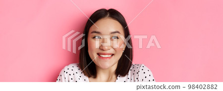 Beauty and lifestyle concept. Headshot of excited asian girl smiling, looking left at promo banner, standing over pink background Beauty and lifestyle concept. Headshot of excited asian girl smiling, looking left at promo banner, standing over pink background 98402882