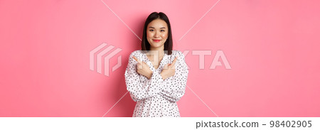 Beautiful asian girl in romantic dress pointing fingers sideways, showing two variants on shopping, smiling at camera, standing against pink background 98402905