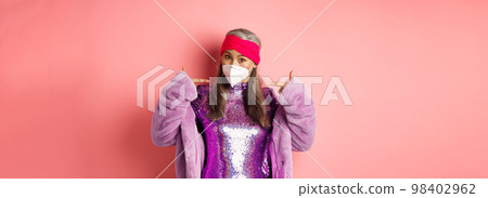 Covid-19, pandemic and fashion concept. Fashionable asian grandmother wearing glittering party dress and respirator, pointing at mask, pink background 98402962