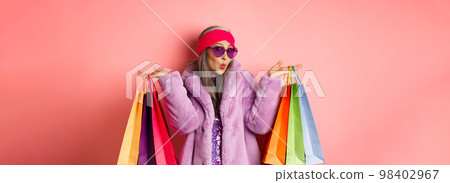Stylish asian senior woman going shopping, wearing trendy clothes and sunglasses, holding store bags with clothes and purchases, pink background Stylish asian senior woman going shopping, wearing trendy clothes and sunglasses, holding store bags with clothes and purchases, pink background 98402967