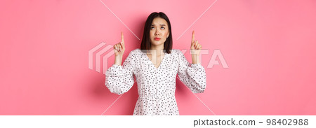 Worried and concerned asian girl in cute dress frowning, pointing and looking up with sad face, standing over pink background Worried and concerned asian girl in cute dress frowning, pointing and looking up with sad face, standing over pink background 98402988