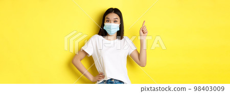 Covid, health care and pandemic concept. Asian female model in medical mask and white t-shirt pointing finger at upper left corner logo, showing promotion, yellow background Covid, health care and pandemic concept. Asian female model in medical mask and white t-shirt pointing finger at upper left corner logo, showing promotion, yellow background 98403009