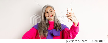 Beautiful and stylish asian senior woman taking selfie on smartphone, smiling and posing in trendy pink sweater, standing over white background 98403013