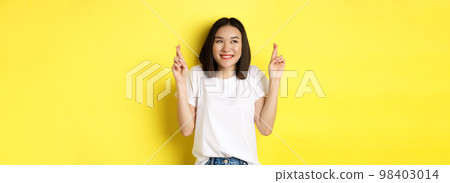 Hopeful asian girl smiling optimistic, feeling lucky, cross fingers and making wish, standing over yellow background 98403014