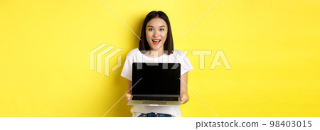 Young asian woman demonstrate online offer, showing blank laptop screen and smiling, standing over yellow background Young asian woman demonstrate online offer, showing blank laptop screen and smiling, standing over yellow background 98403015