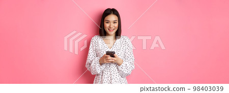 Beautiful asian lady reading message and smiling, using mobile phone, standing in cute dress against pink background Beautiful asian lady reading message and smiling, using mobile phone, standing in cute dress against pink background 98403039