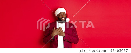 Christmas, party and holidays concept. Grateful african american man in santa hat saying thank you, holding hands on heart and smiling, feeling touched, standing against red background 98403087