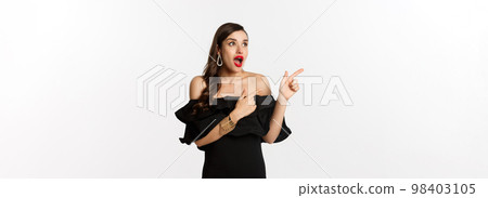 Fashion and beauty. Amazed beautiful woman in black dress open mouth fascinated, pointing fingers and looking left, white background 98403105