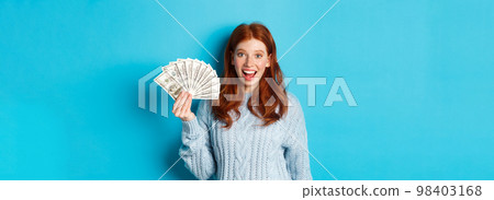 Happy redhead girl in sweater, staring excited at camera, showing money dollars, standing against blue background 98403168