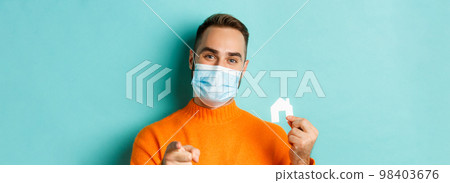 Real estate and coronavirus pandemic concept. Close-up of adult man in medical mask pointing at you and showing small paper house, standing over light blue background 98403676