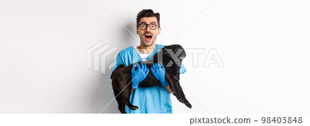 Vet clinic concept. Happy male doctor veterinarian holding cute black pug dog, staring at camera amazed, white background 98403848