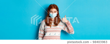 Winter, coronavirus and social distancing concept. Impressed redhead girl in face mask, showing thumb up in approval, like and praise, standing over blue background 98403963