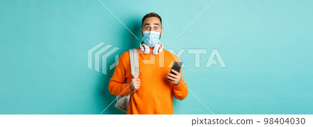 Young man in face mask using mobile phone, holding backpack, staring impressed at camera, standing against light blue background Young man in face mask using mobile phone, holding backpack, staring impressed at camera, standing against light blue background 98404030