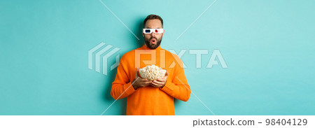 Handsome man in orange sweater and 3d glasses watching movies thrilled, holding popcorn, looking amazed, standing over blue background 98404129