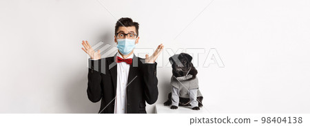 Coronavirus, pets and celebration concept. Amazed young man in face mask and suit staring at camera surprised, cute black dog sitting near owner in party outfit, white background 98404138