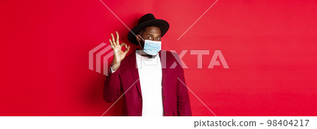 Covid-19 and fashion concept. Stylish african american man in hat and blazer, wearing face mask and showing okay sign, standing over red background 98404217