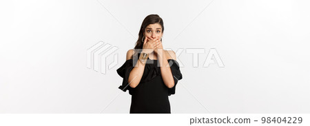 Fashion and beauty. Attractive woman in black dress cover mouth and gasping shocked, staring at camera worried, standing over white background Fashion and beauty. Attractive woman in black dress cover mouth and gasping shocked, staring at camera worried, standing over white background 98404229