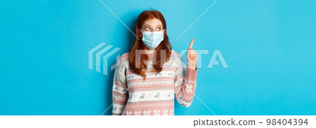 Winter, covid-19 and quarantine concept. Curious redhead girl in medical mask picking product, looking and pointing at upper left corner promo, blue background Winter, covid-19 and quarantine concept. Curious redhead girl in medical mask picking product, looking and pointing at upper left corner promo, blue background 98404394