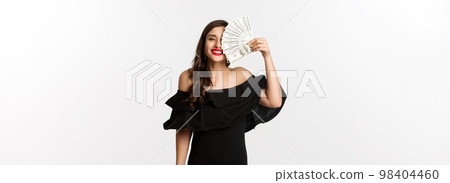 Fashion and shopping concept. Happy young woman in black dress, with red lips, holding money and smiling satisfied, standing over white background 98404460
