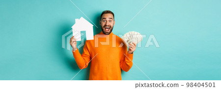 Real estate concept and mortgage concept. Excited man showing dollars and paper maket of house, buying flat, standing over blue background 98404501