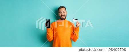 Online shopping. Thoughtful man shop in internet, showing mobile screen and credit card, looking at upper left corner and thinking, standing over light blue background 98404798