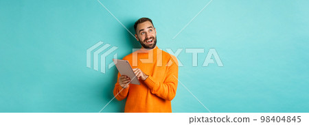 Handsome man thinking and using digital tablet, shopping online and pondering ideas, looking at upper left corner, standing over light blue background Handsome man thinking and using digital tablet, shopping online and pondering ideas, looking at upper left corner, standing over light blue background 98404845