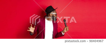 Passionate black male singer performing against red background, singing into microphone, wearing party outfit, standing over red background 98404967