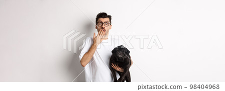Image of handsome young man holding cute dog and gasping surprised. Pet owner staring at camera shocked, carry black pug in arm, white background 98404968