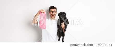 Image of hipster guy pet owner, holding cute black pug and dog poop bag, standing over white background 98404974